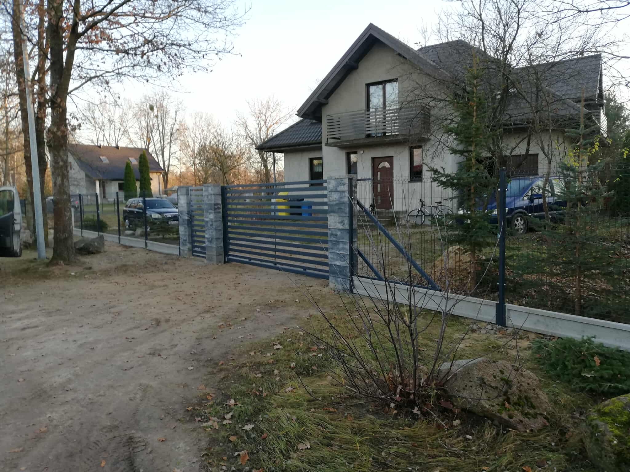 Nowoczesna, pozioma brama wjazdowa z szarego metalu i kamiennymi słupkami, zamykająca posesję z domem w tle. Widoczny fragment ogrodzenia panelowego.