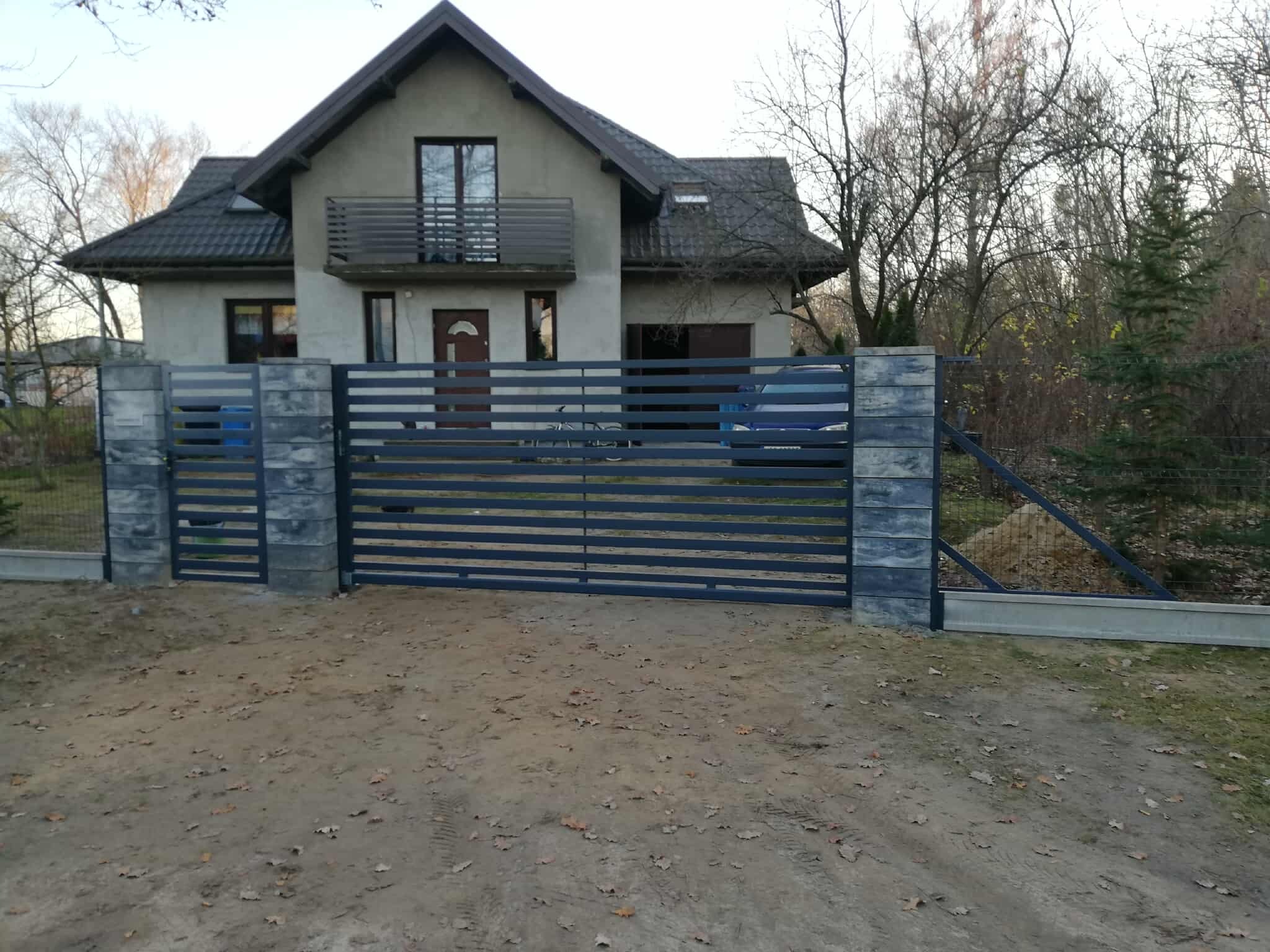Nowoczesna, pozioma brama wjazdowa z szarego metalu i betonowych słupków, na tle domu z balkonem i jesiennego ogrodu. Widok z zewnątrz posesji, w pochmurny dzień.
