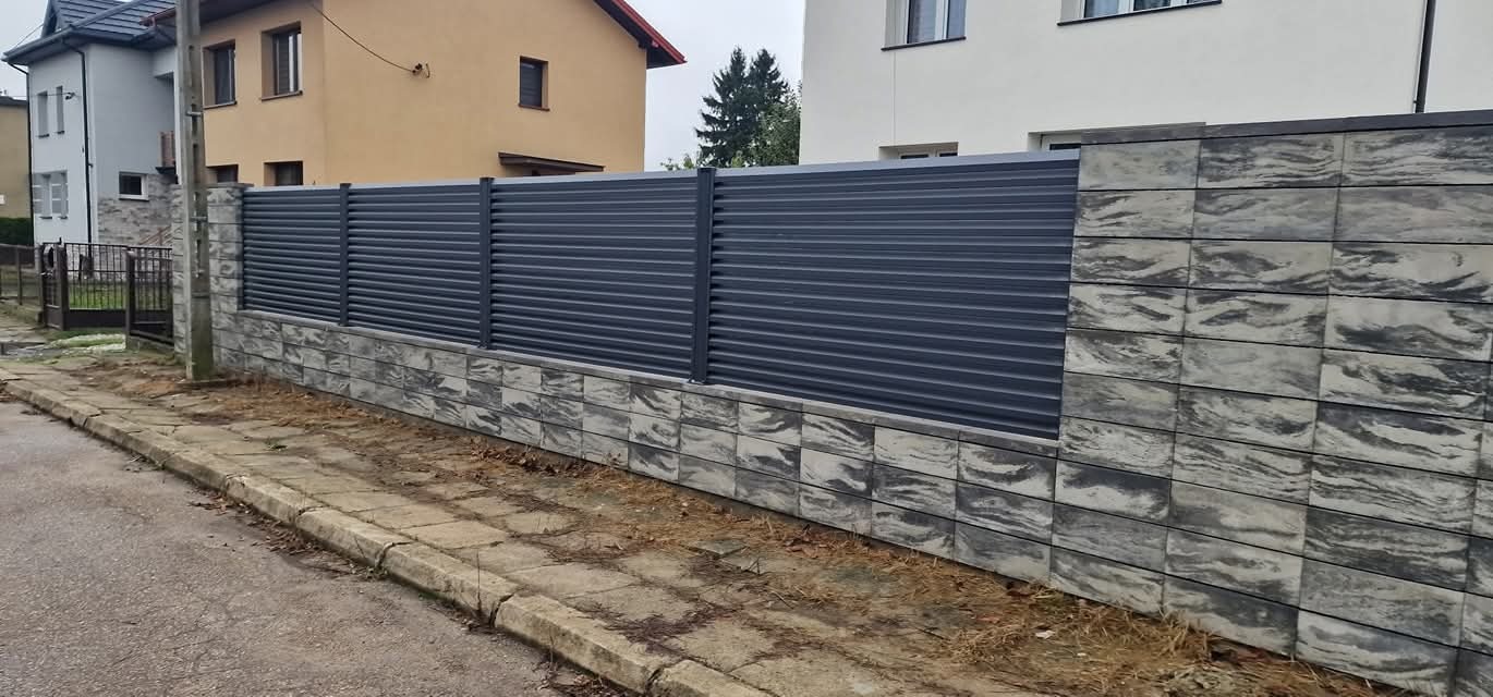 Nowoczesne ogrodzenie panelowe w kolorze grafitowym, z podmurówką z szarej kostki imitującej kamień, na tle zabudowy jednorodzinnej.