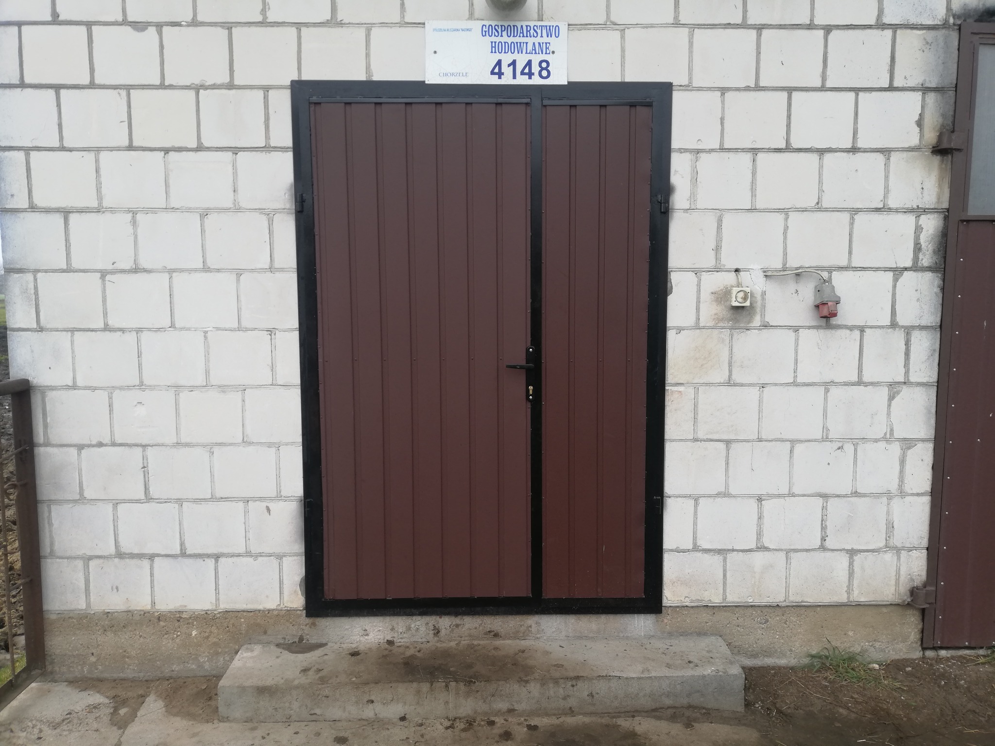 Brązowa brama dwuskrzydłowa z czarną ramą, zamontowana w ścianie z białych bloczków. Nad bramą tabliczka z adresem 'Gospodarstwo Hodowlane 4148'.