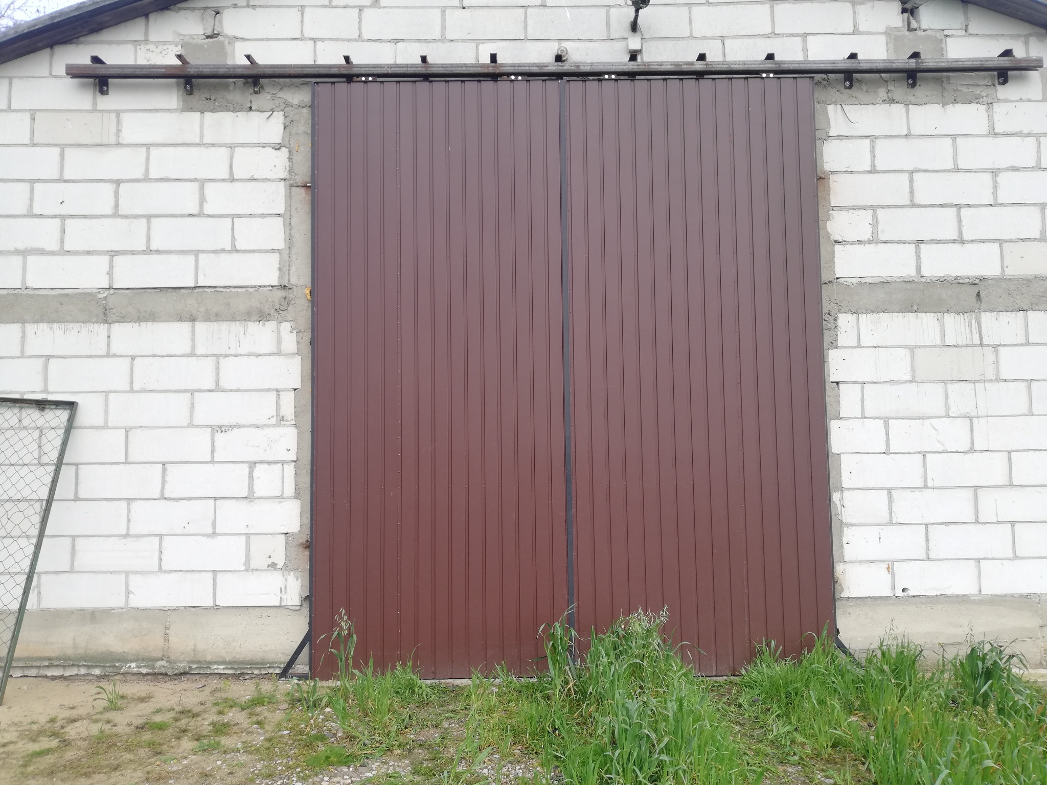 Brązowa, przesuwna brama garażowa z pionowymi panelami, zamontowana na białym murze z bloczków. Widoczne metalowe prowadnice i fragment siatki ogrodzeniowej po lewej.