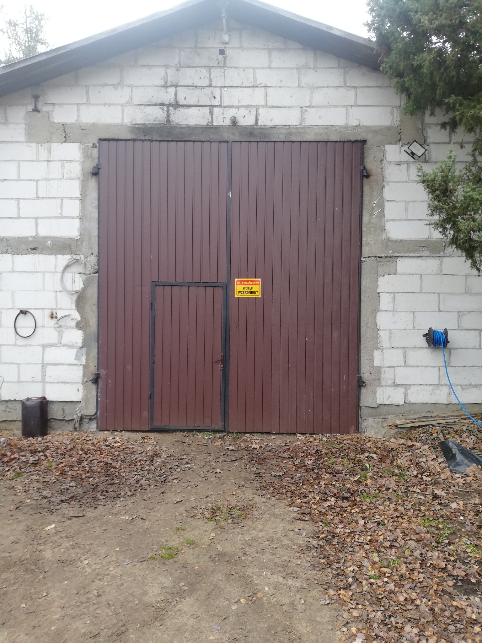 Brązowa brama garażowa z małym wejściem, umieszczona w budynku z białych bloczków. Na bramie żółta tabliczka ostrzegawcza. Obok bębnowy zwijak z niebieskim wężem.
