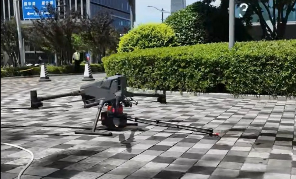 Dron z lancą do mycia powierzchni na zewnątrz, na tle zieleni i budynków. Widoczne ślady wody na kostce brukowej.