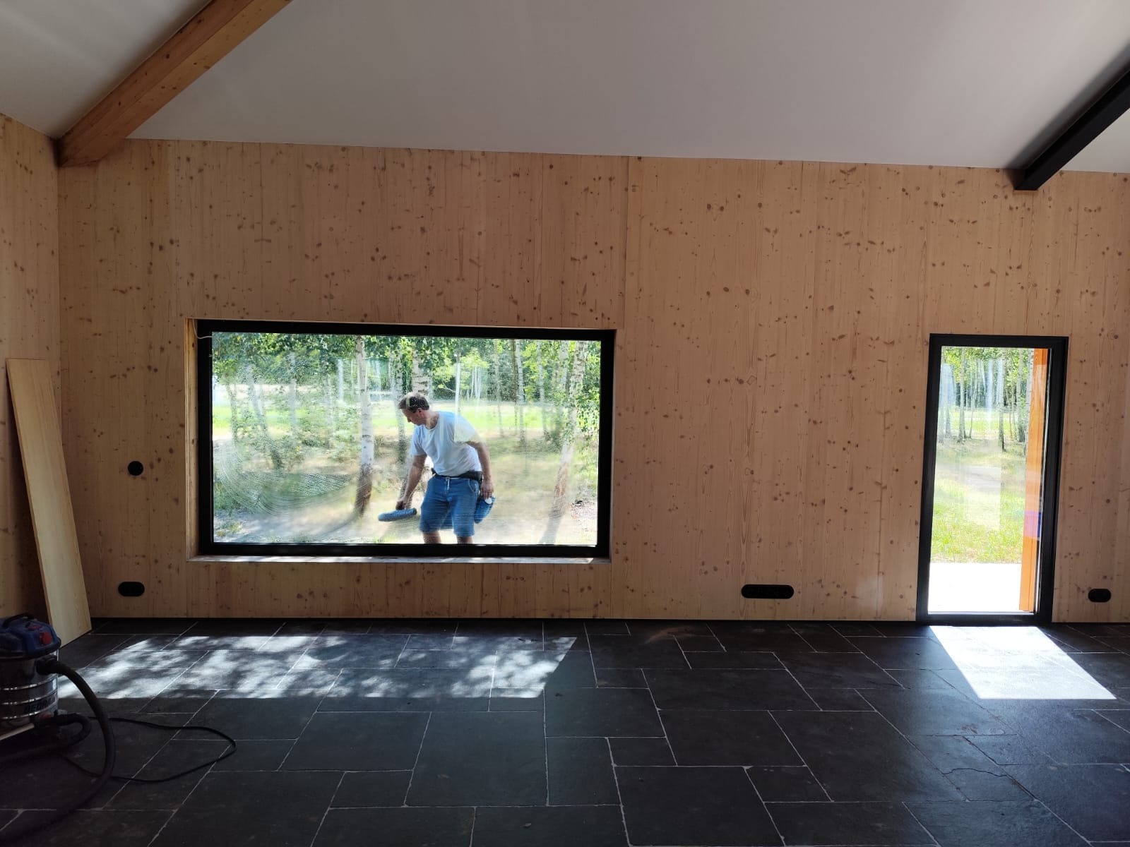 Mężczyzna myje okno w drewnianym domu z widokiem na las. Wnętrze w trakcie wykańczania, widoczna podłoga z ciemnych płytek i drewniane ściany.