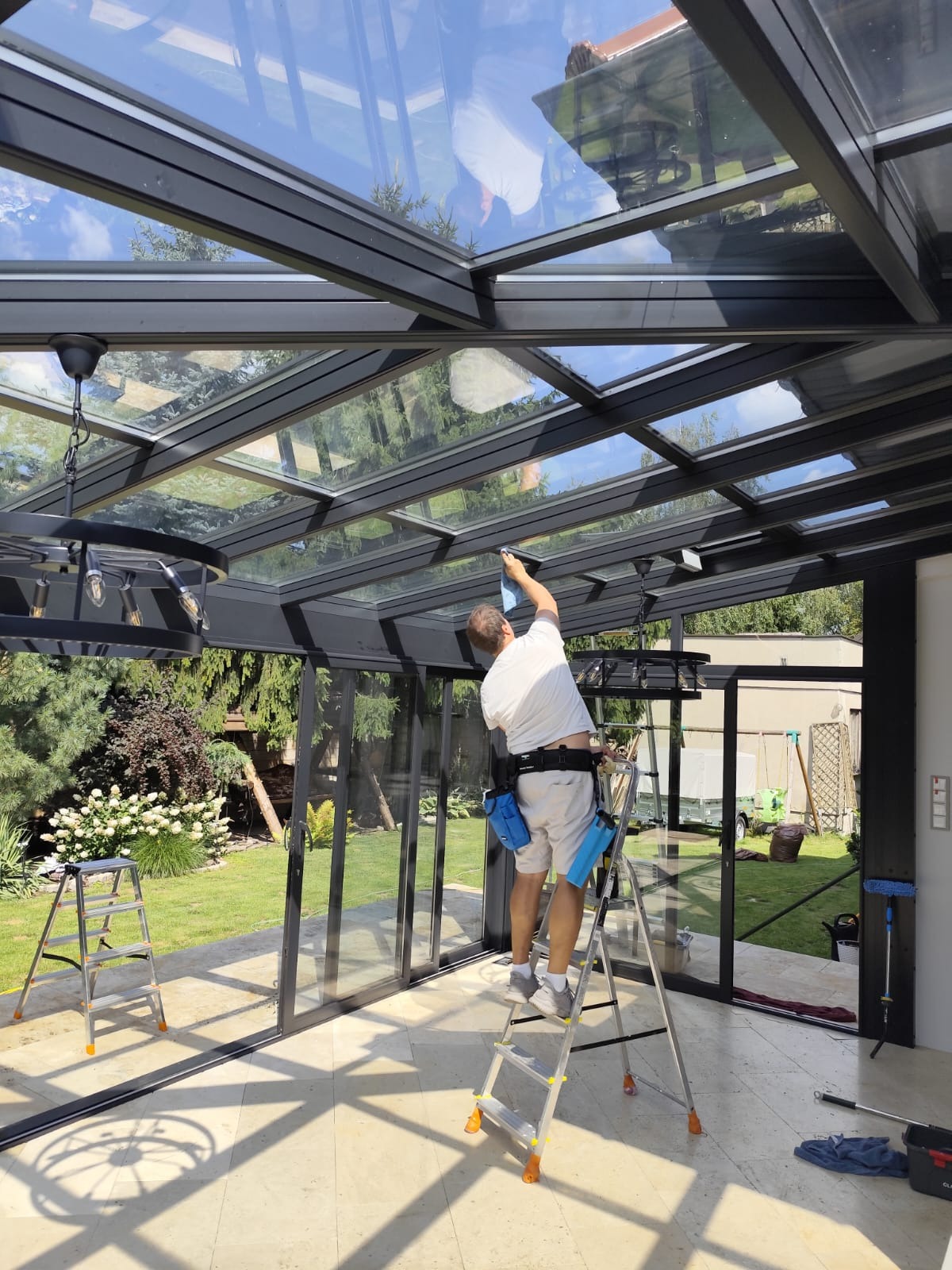Pracownik na drabinie czyści szklany dach oranżerii. Widoczne narzędzia, otoczenie ogrodu i elementy konstrukcyjne dachu. Jasne światło słoneczne.
