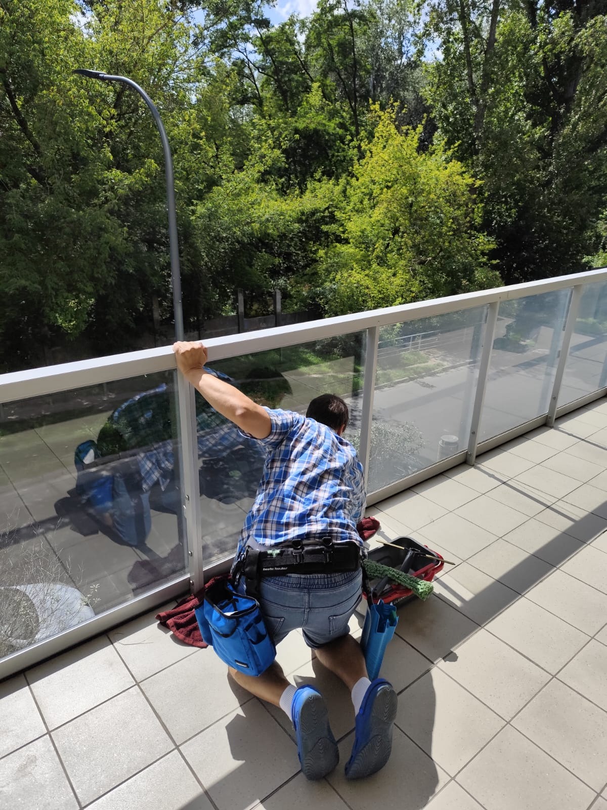 Mężczyzna w niebieskiej koszuli w kratę klęczy na balkonie, myjąc szklane panele balustrady. Widoczne narzędzia i akcesoria do mycia okien. W tle zieleń drzew.