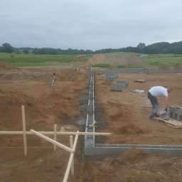 Damian Jabłonowski - Budowa fundamentów z bloczków betonowych na placu budowy. Widoczny pracownik układający bloczki, poziomica laserowa i drewniane szalunki. Tło: pole i pochmurne niebo.