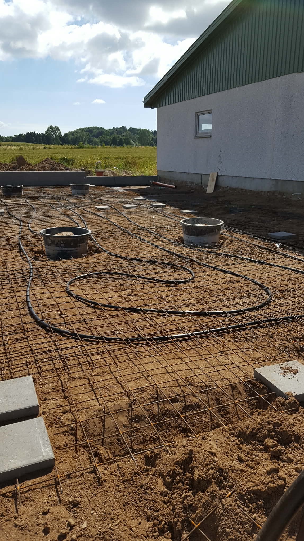 Przygotowanie podłoża pod kostkę brukową: siatka zbrojeniowa, rury grzewcze i betonowe obrzeża na piasku. W tle budynek i pole uprawne.