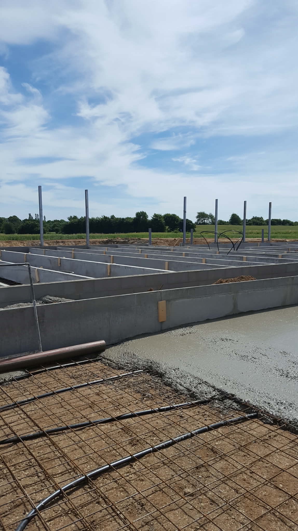 Zalewanie betonem fundamentów pod budynek. Widoczna siatka zbrojeniowa i szalunki. W tle zieleń drzew i błękitne niebo z chmurami.