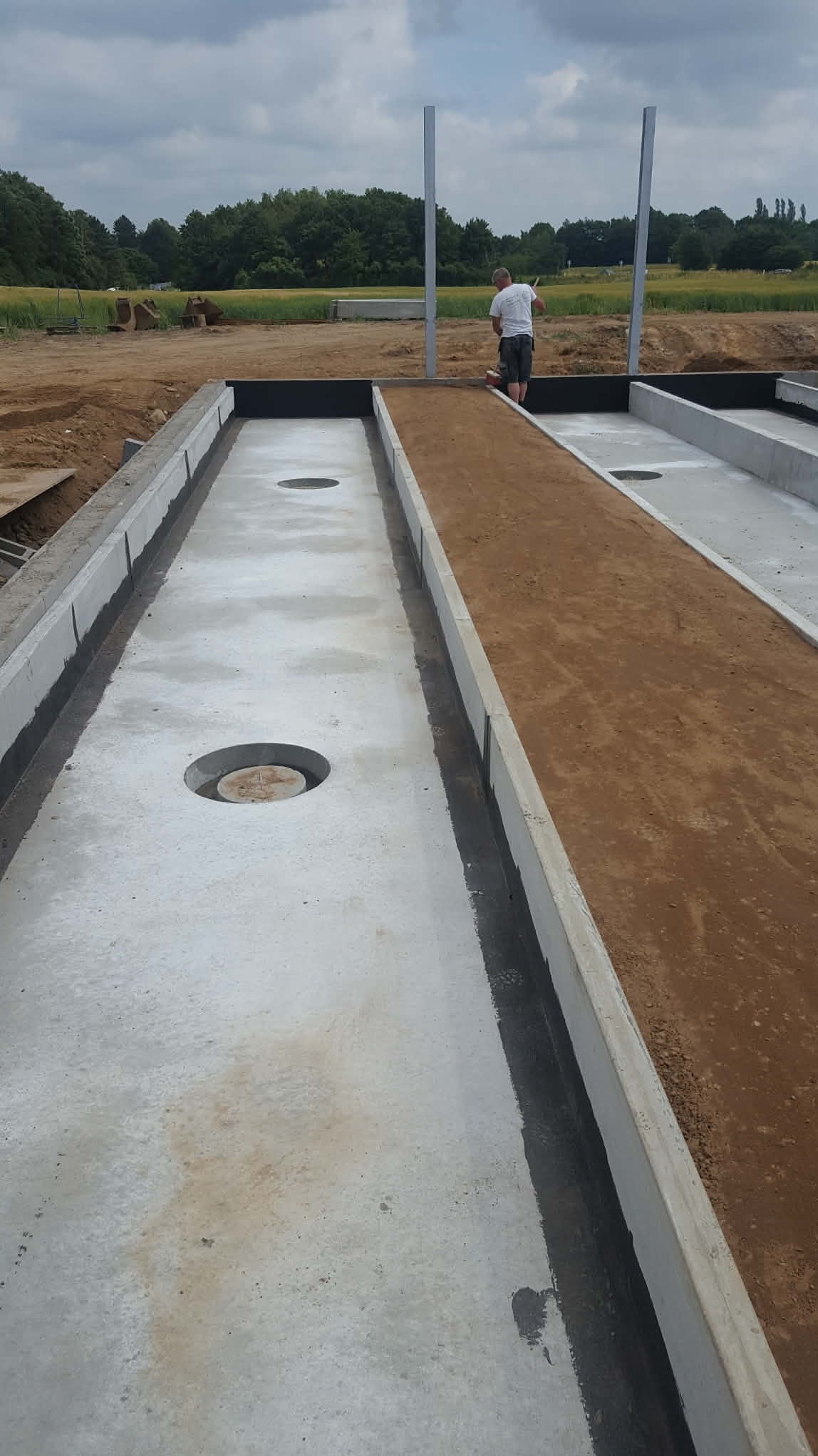 Betonowe koryta z ziemią i otworami, prawdopodobnie element drenażu lub fundamentów. W tle osoba przy pracy i zielone pola. Widok z góry.