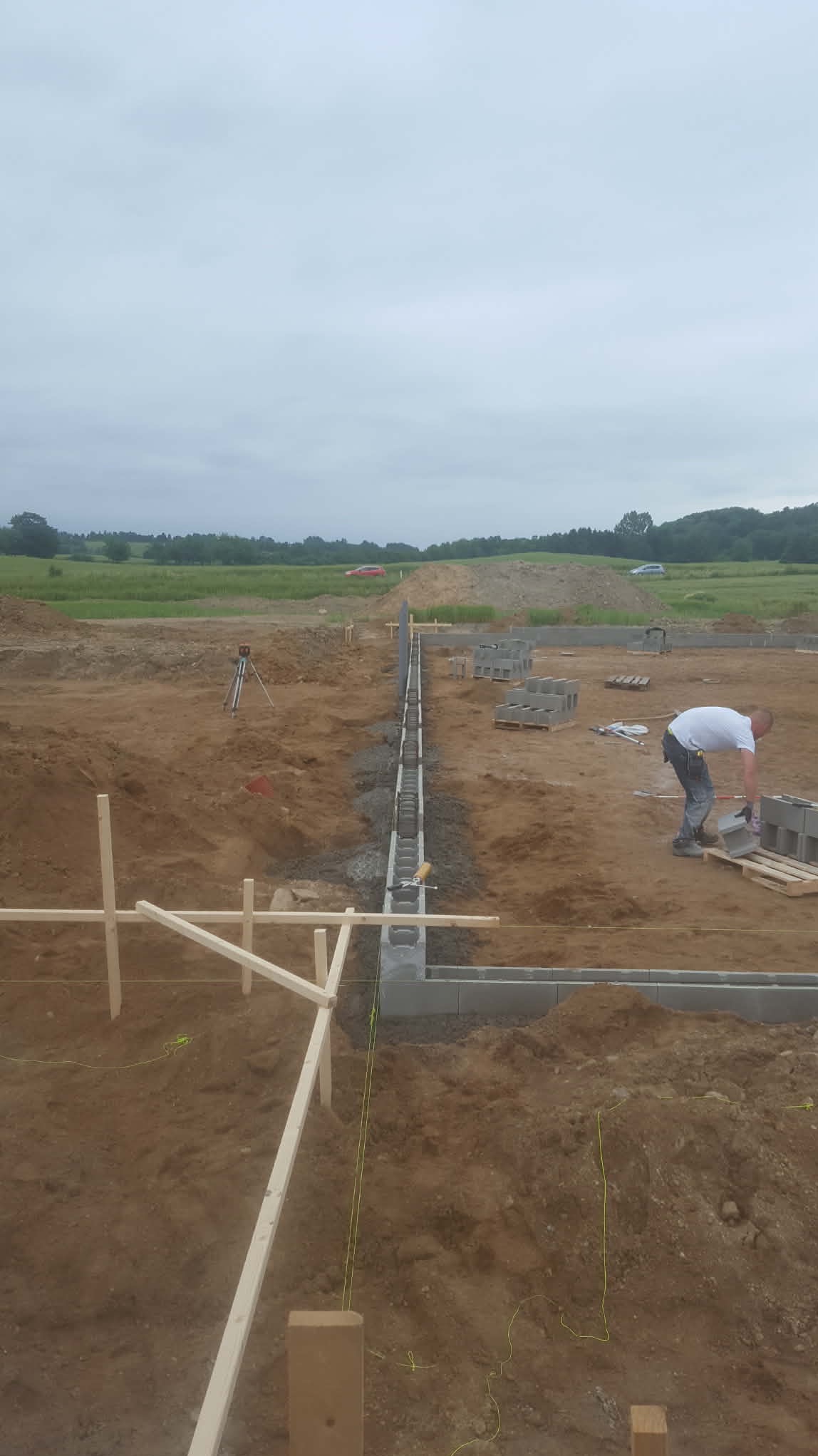 Budowa fundamentów z bloczków betonowych na placu budowy. Widoczny pracownik układający bloczki, poziomica laserowa i drewniane szalunki. Tło: pole i pochmurne niebo.