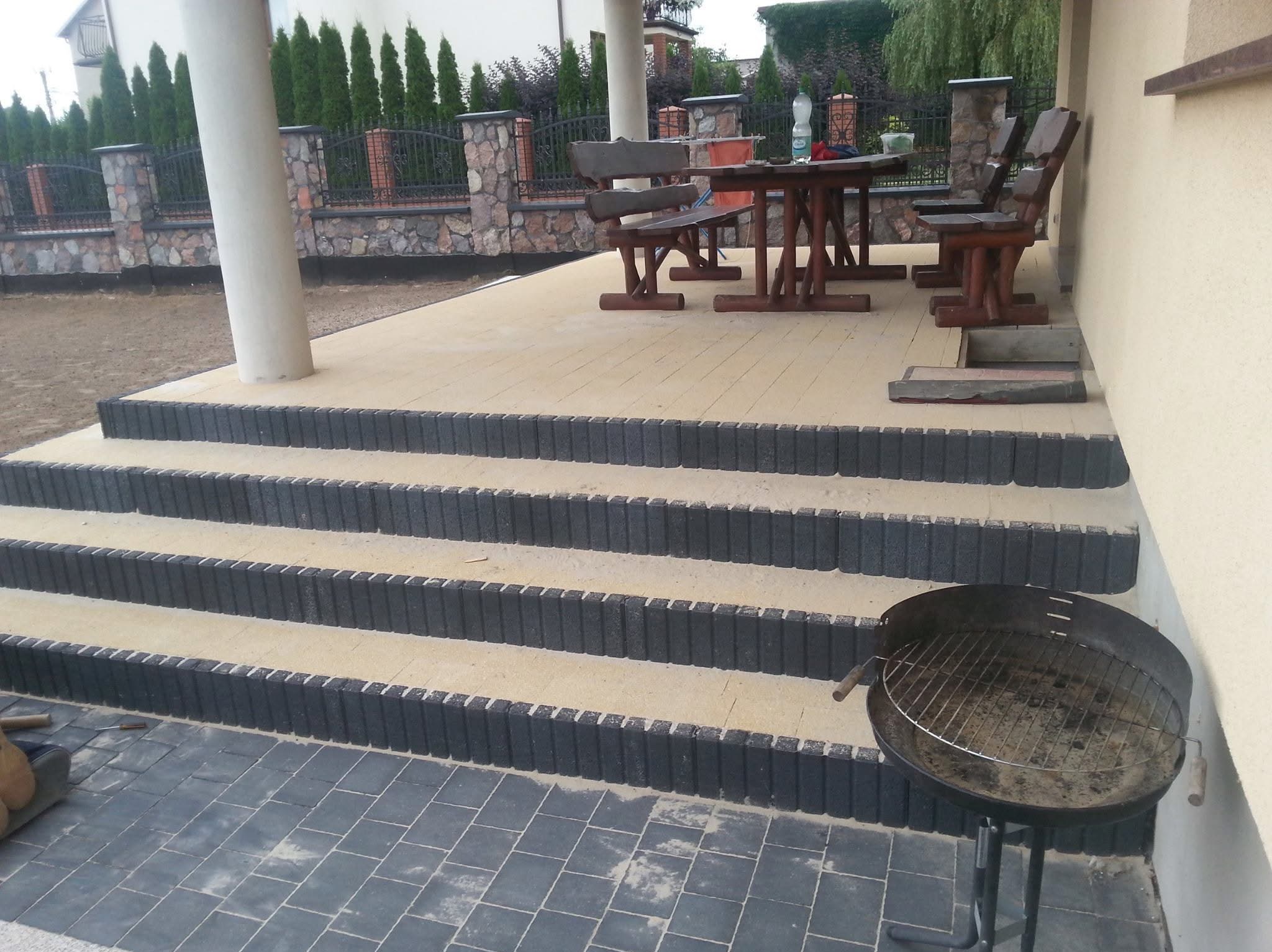 Ułożona kostka brukowa tworząca taras z schodami. Widoczne meble ogrodowe i grill. W tle ogrodzenie z kamienia i metalu, oraz wysokie tuje.