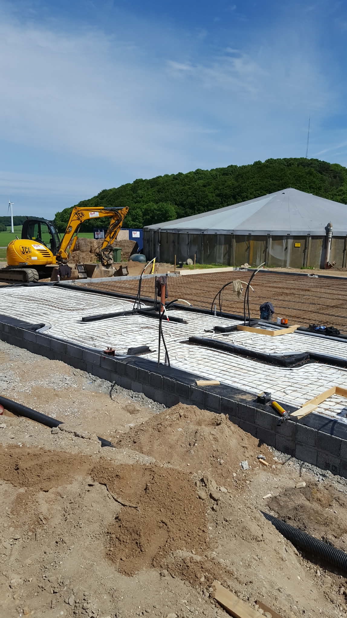 Przygotowanie podłoża pod budynek z użyciem mini koparki JCB, widoczne fundamenty z bloczków betonowych i zbrojenie. W tle zielone wzgórze i budynek gospodarczy.