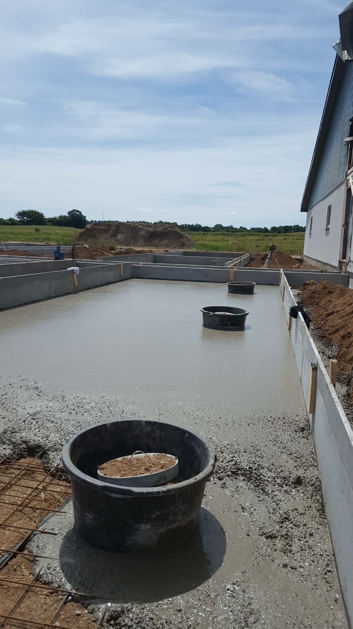 Zalewanie fundamentów w Ostródzie. Widoczne szalunki, świeży beton i wiaderka na powierzchni. W tle zielone pola i błękitne niebo. Prace budowlane w toku.