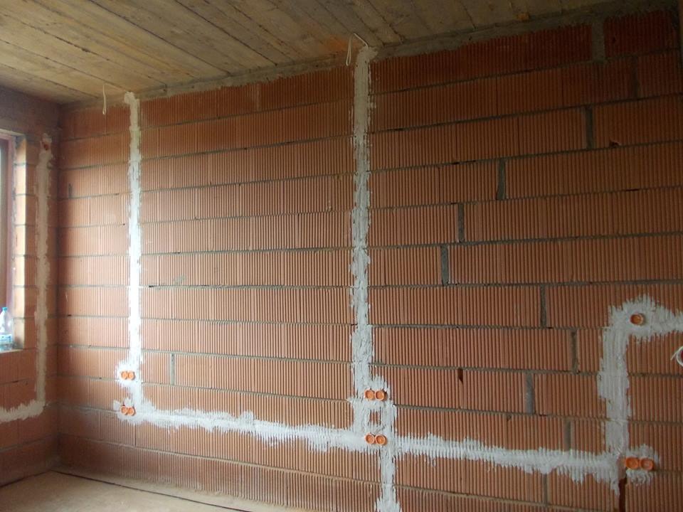 Ściana z czerwonej cegły z wyżłobionymi i wypełnionymi gipsem bruzdami pod instalację elektryczną. Widoczne puszki pod gniazdka.