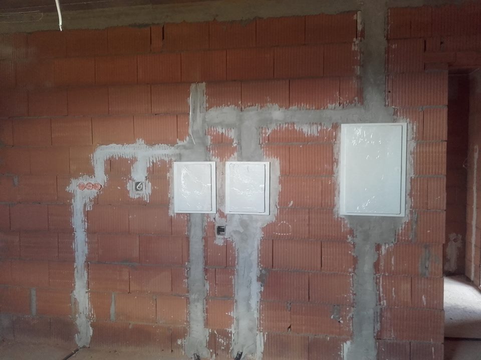 Murowana ściana z trzema białymi skrzynkami i bruzdami wypełnionymi gipsem, prawdopodobnie pod kable. Widoczne gniazdka i przewody. Surowy stan wykończenia.