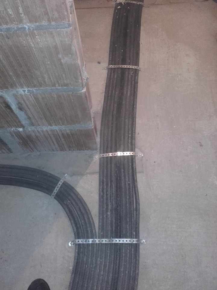 Wiązka przewodów elektrycznych w peszlu, przymocowana metalowymi obejmami do ściany i podłoża. Widoczne kable ułożone w łuk i prosto, przed zalaniem posadzki.