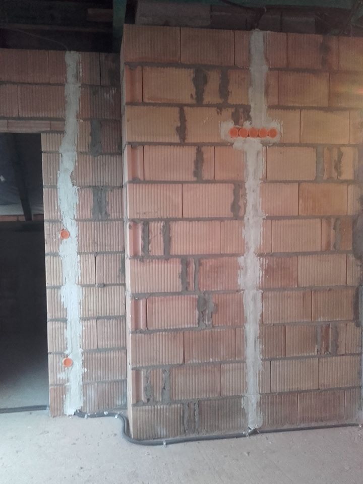 Ceglana ściana w trakcie prac instalacyjnych: widoczne bruzdy wypełnione zaprawą, pomarańczowe puszki elektryczne i przewody przy podłodze. Surowy stan wykończenia.