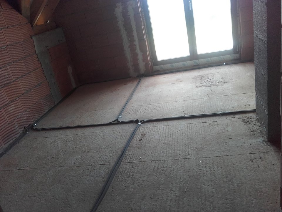 Kable elektryczne ułożone krzyżowo na podłodze w surowym stanie, przed wylaniem posadzki. Widoczne ściany z cegły i okno w tle. Kable przymocowane do podłoża.