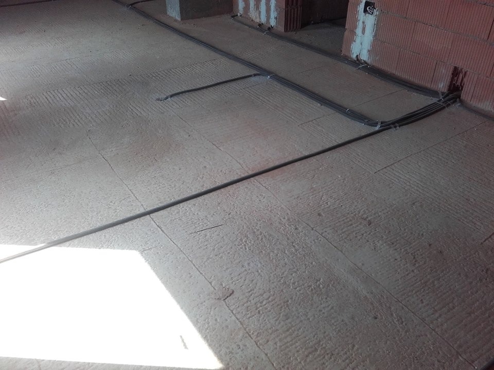 Kable elektryczne ułożone na podłodze w surowym stanie, przed wylaniem posadzki. Widoczne ściany z czerwonej cegły i puszki elektryczne.