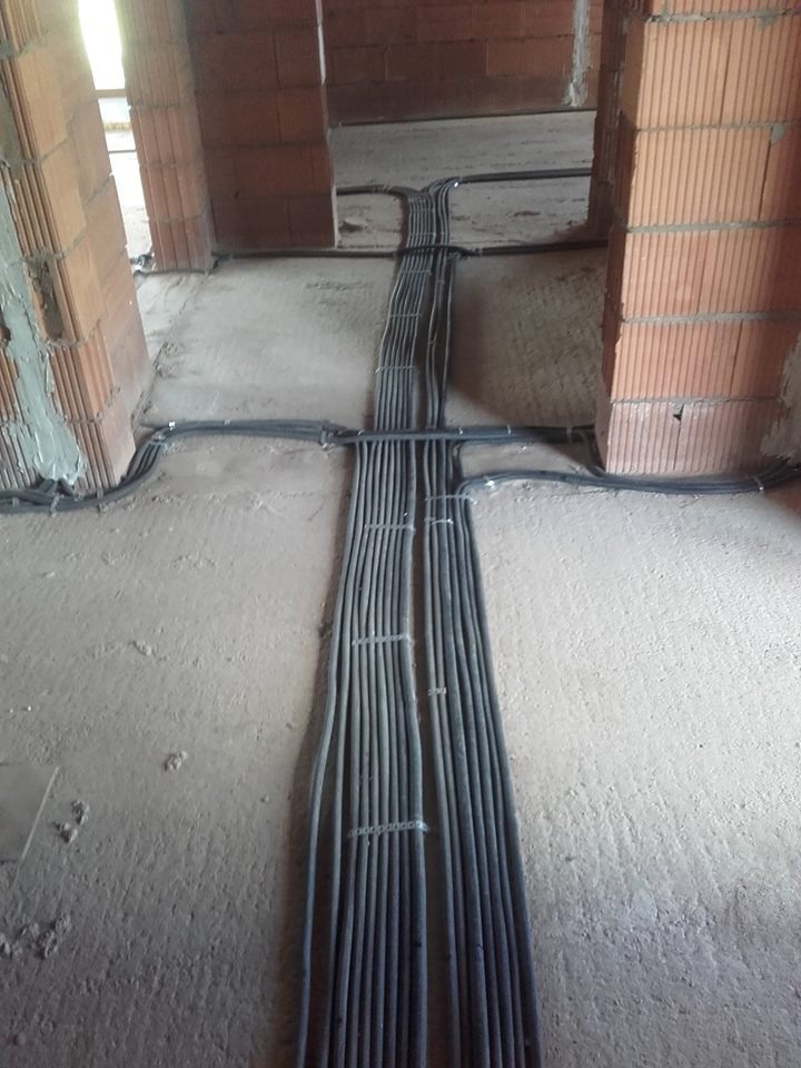 Szereg szarych kabli elektrycznych ułożonych równolegle na surowym betonowym podłożu wewnątrz budynku w trakcie budowy, widoczne ceglane ściany.