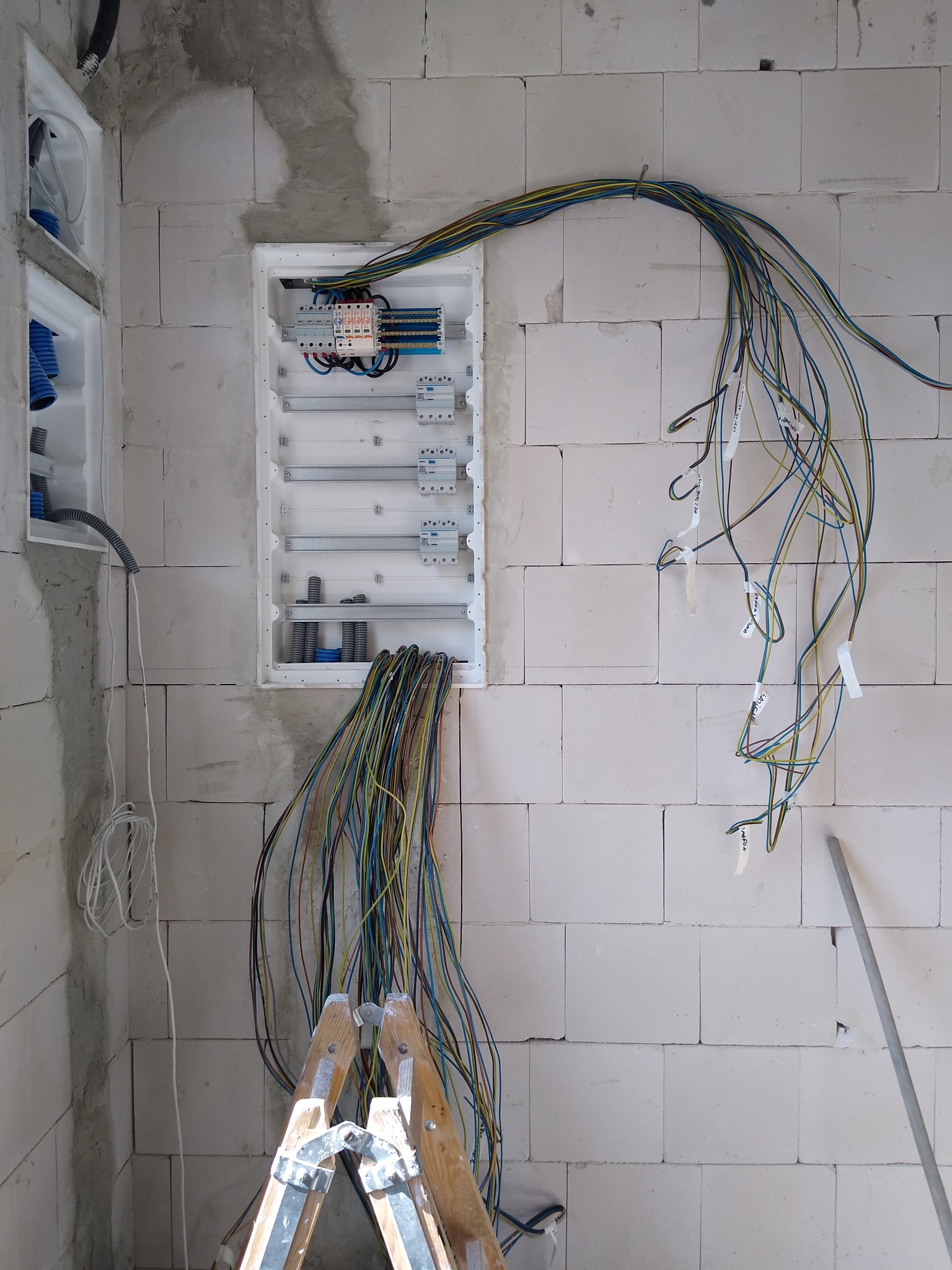 Rozdzielnia elektryczna w trakcie montażu w surowym wnętrzu. Widoczne kable, drabina i ściana z betonu komórkowego. Etap prac instalacyjnych.