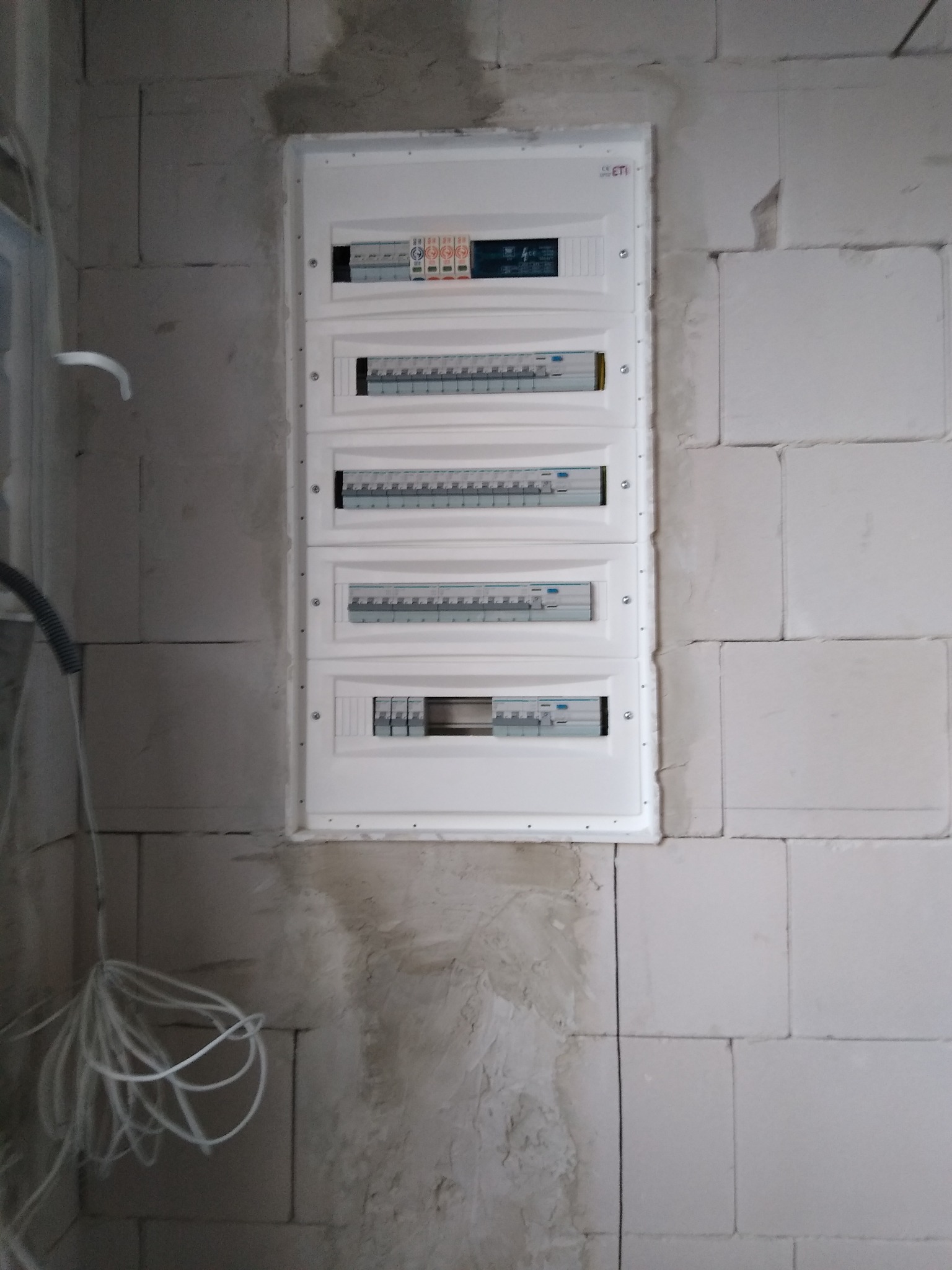 Rozdzielnica elektryczna, biała, zamontowana na surowej ścianie z betonu komórkowego. Widoczne zabezpieczenia i przewody.