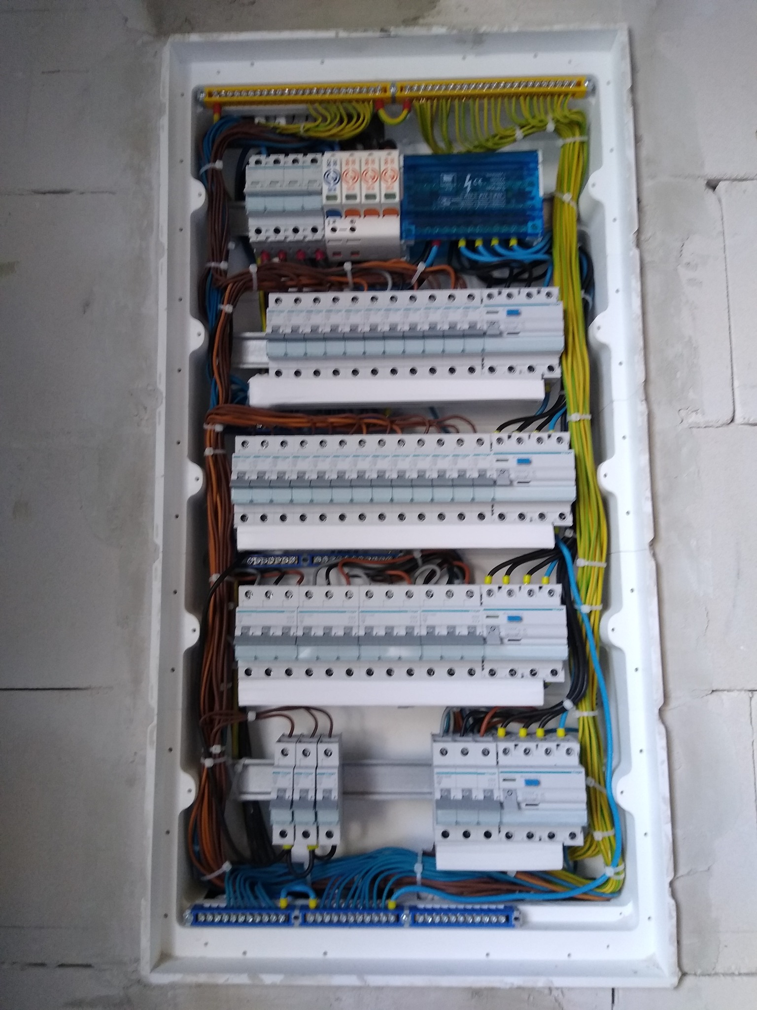 Rozdzielnica elektryczna wnękowa z bezpiecznikami, przewodami w kolorach: żółtym, niebieskim, brązowym i czarnym. Widoczne elementy montażowe i ściana z betonu komórkowego.