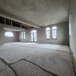 Łukasz Madeński - Surowe wnętrze z gładkimi ścianami i sufitem po tynkowaniu maszynowym. Widoczne okna łukowe i kable na podłodze. Stan surowy zamknięty przed wykończeniem.