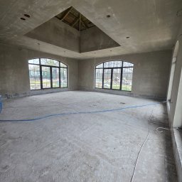 Łukasz Madeński - Surowe wnętrze po tynkowaniu maszynowym: szare ściany, okna z czarnymi ramami, widoczne instalacje, sufit z otworem na poddasze. Wnętrze w stanie deweloperskim.
