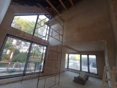 Wnętrze domu w trakcie tynkowania maszynowego. Widoczne rusztowanie, duże okna i surowe ściany pokryte tynkiem. Charakterystyczne skośne okno na górze.