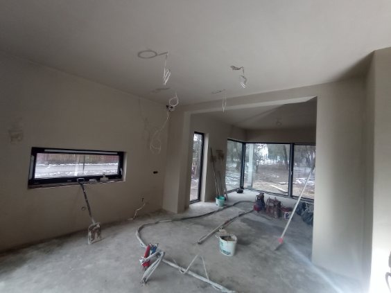 Wnętrze po nałożeniu tynków: gładkie, jasne ściany, widoczne przewody elektryczne, okno z widokiem na zewnątrz, sprzęt do tynkowania na podłodze. Minimalistyczny design.