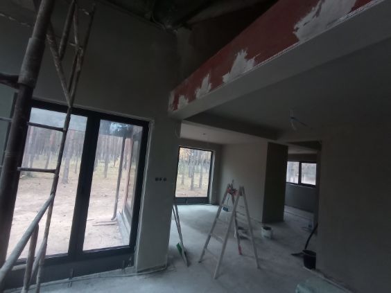 Wnętrze domu w trakcie prac wykończeniowych: świeżo otynkowane ściany w szarym odcieniu, drabiny, duże okna z widokiem na las. Etap przygotowania do dalszych prac.