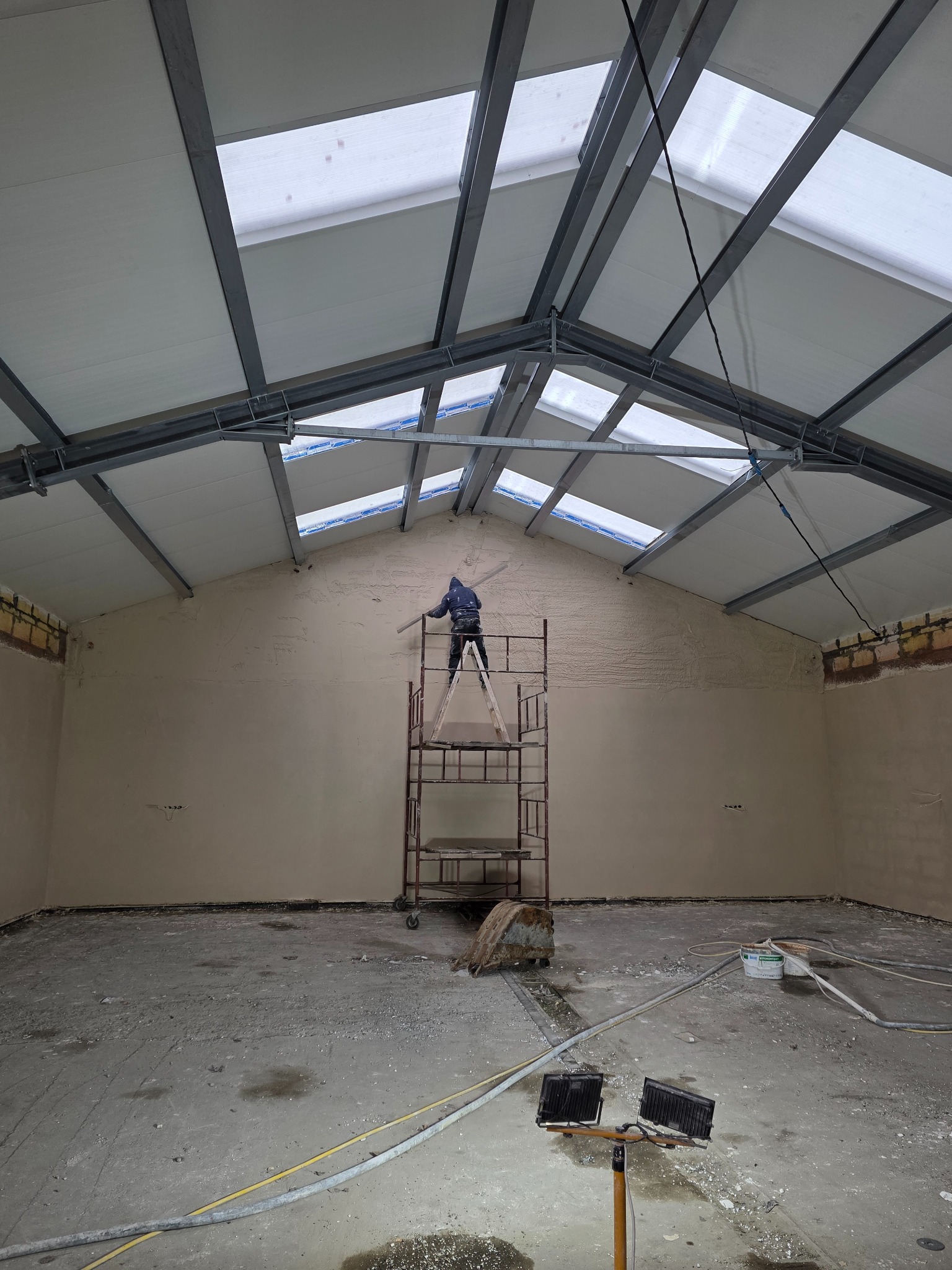 Tynkowanie ścian wewnątrz hali z oknami dachowymi. Pracownik na rusztowaniu aplikuje tynk. Widoczne narzędzia i oświetlenie budowlane.