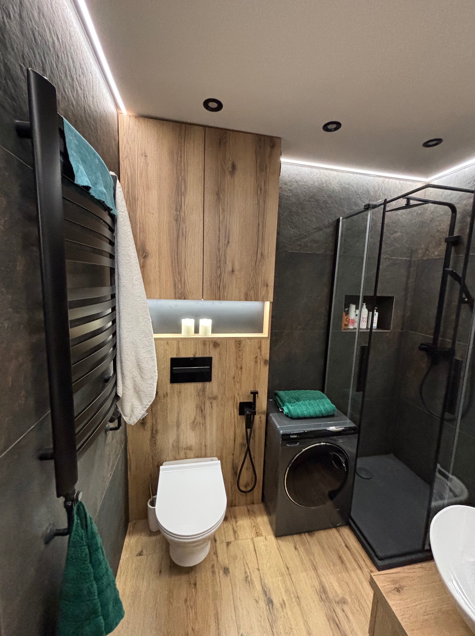 Nowoczesna łazienka z kabiną prysznicową typu walk-in, pralką i drewnianymi akcentami. Oświetlenie LED podkreśla minimalistyczny design i funkcjonalność.