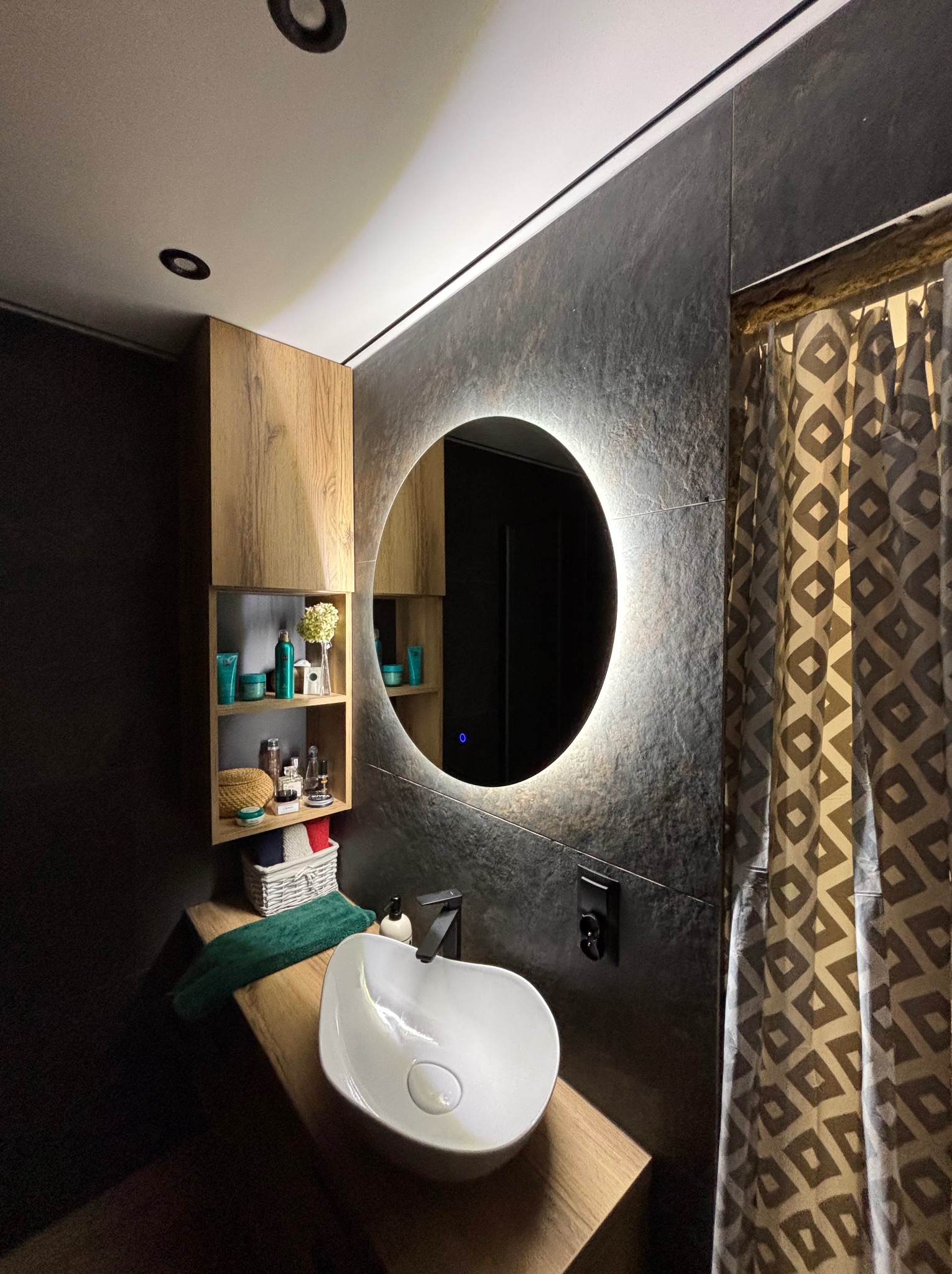 Nowoczesna łazienka z okrągłym lustrem LED, umywalką nablatową i drewnianą szafką. Ściana wykończona ciemnym kamieniem, zasłona w geometryczny wzór.
