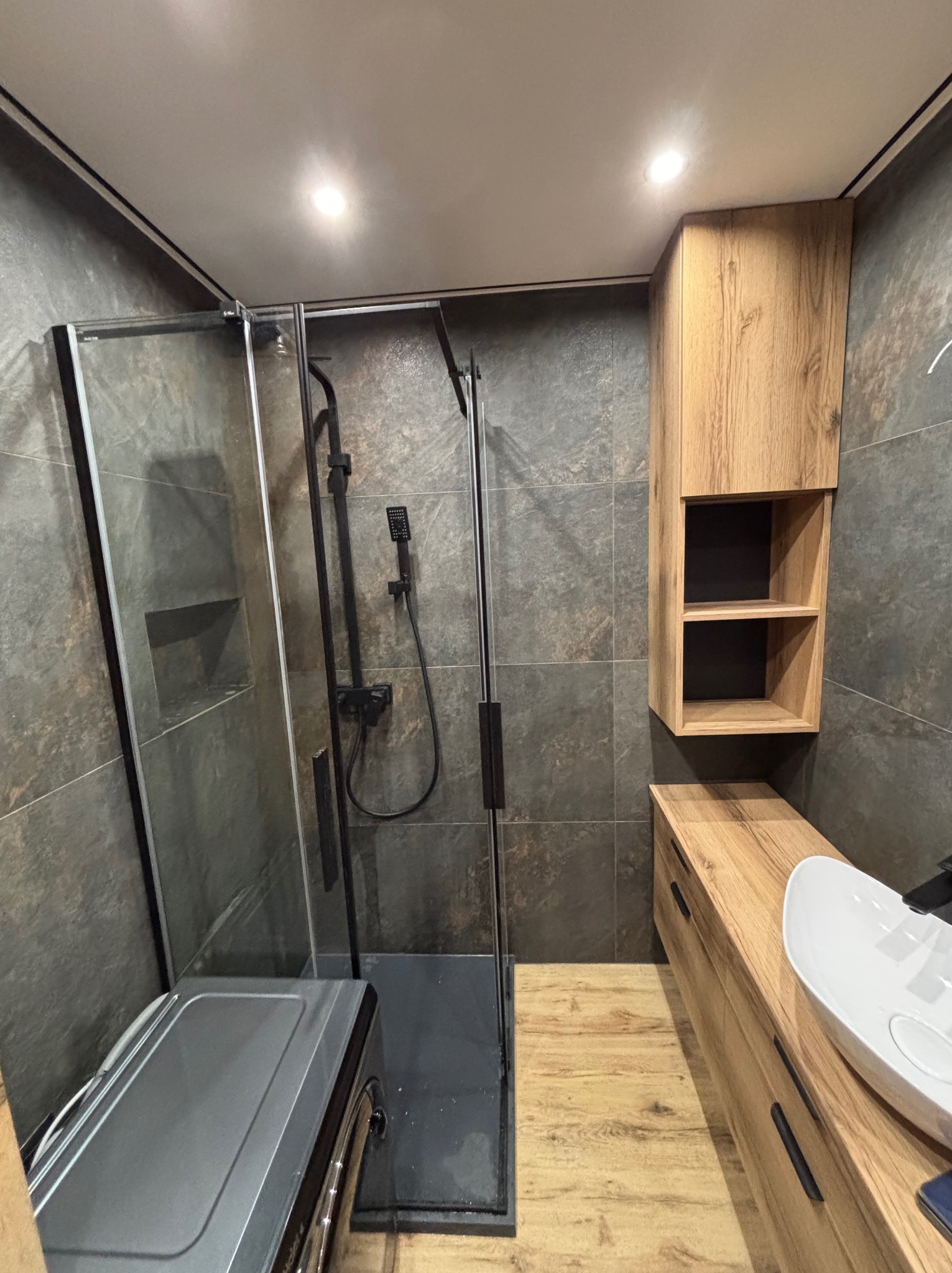 Nowoczesna łazienka z kabiną prysznicową typu walk-in, drewnianą szafką z umywalką nablatową i szarą płytką imitującą kamień na ścianach.
