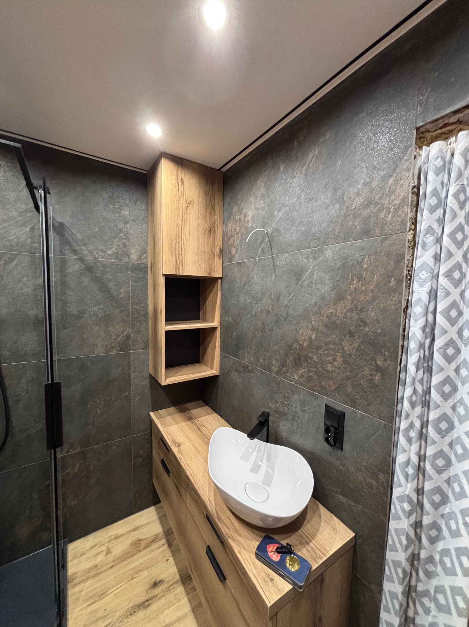 Nowoczesna łazienka z drewnianą szafką pod umywalką i wiszącą szafką. Ciemne płytki na ścianach, biała umywalka nablatowa, czarna armatura i kabina prysznicowa.