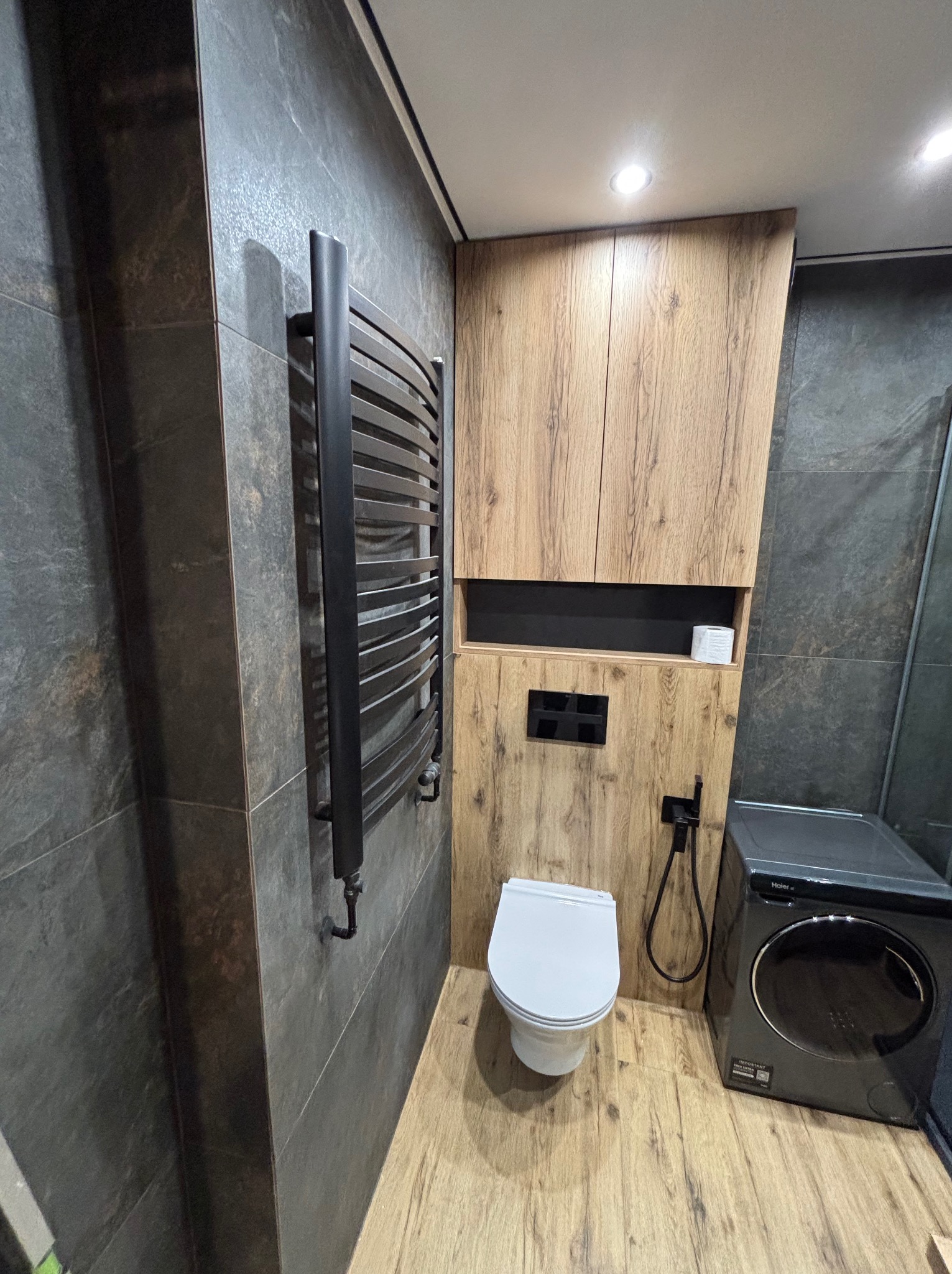 Nowoczesna łazienka z szarymi płytkami, drewnianymi szafkami i grzejnikiem drabinkowym. Uwagę zwraca pralka w ciemnym kolorze oraz minimalistyczna toaleta.