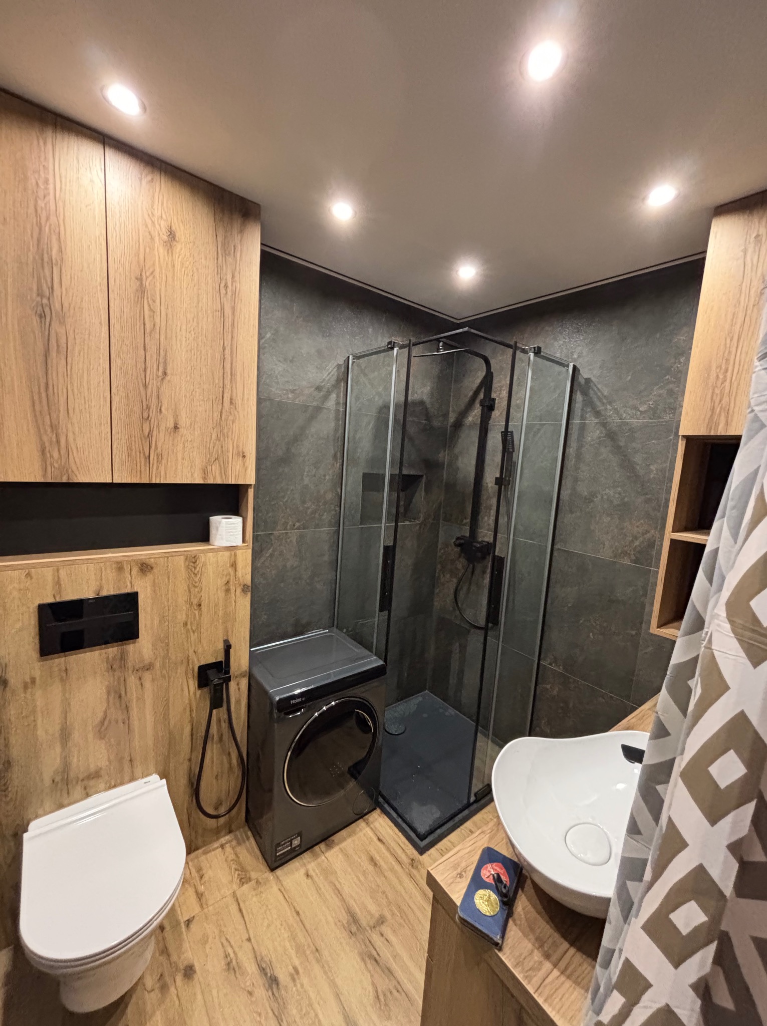 Nowoczesna łazienka z kabiną prysznicową, umywalką nablatową i pralką. Drewnopodobne płytki i szafki tworzą ciepły klimat. Czarna armatura i szara ściana dodają elegancji.