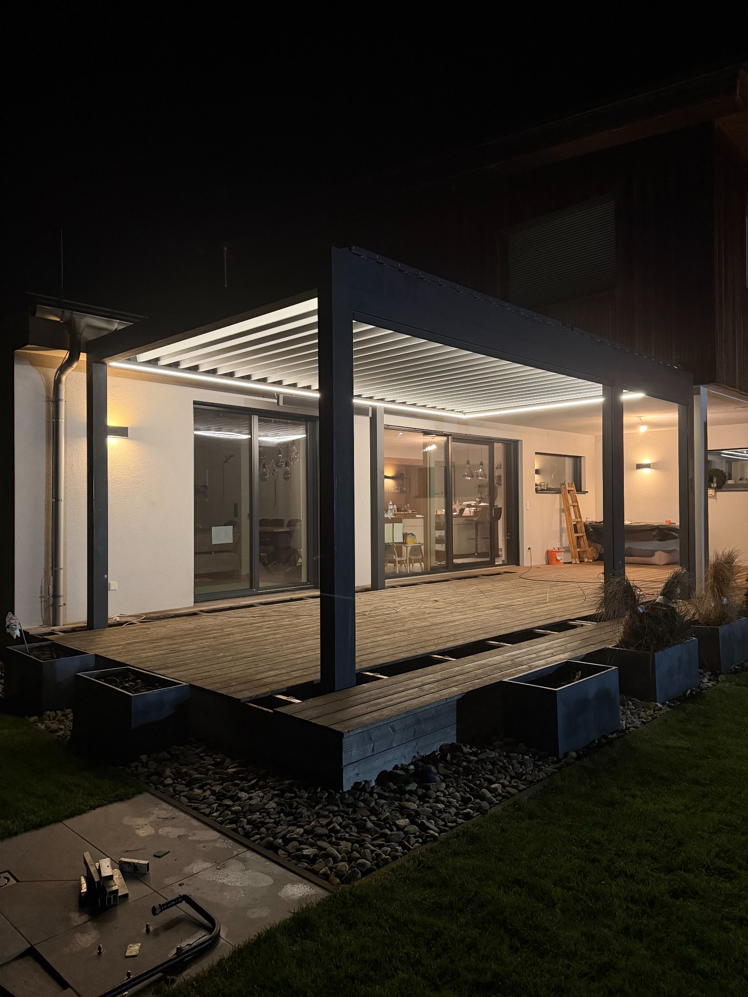 Nowoczesna, oświetlona pergola metalowa z drewnianym tarasem i elementami dekoracyjnymi w ogrodzie, sfotografowana nocą. Widoczne detale konstrukcyjne i oświetlenie LED.