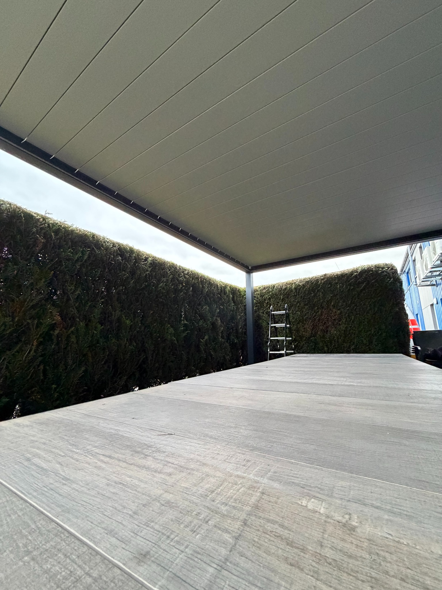 Nowoczesna pergola z szarym zadaszeniem i drewnianą podłogą, otoczona zielonym żywopłotem. Widoczna drabina oparta o żywopłot. Ujęcie z dołu, podkreślające konstrukcję.