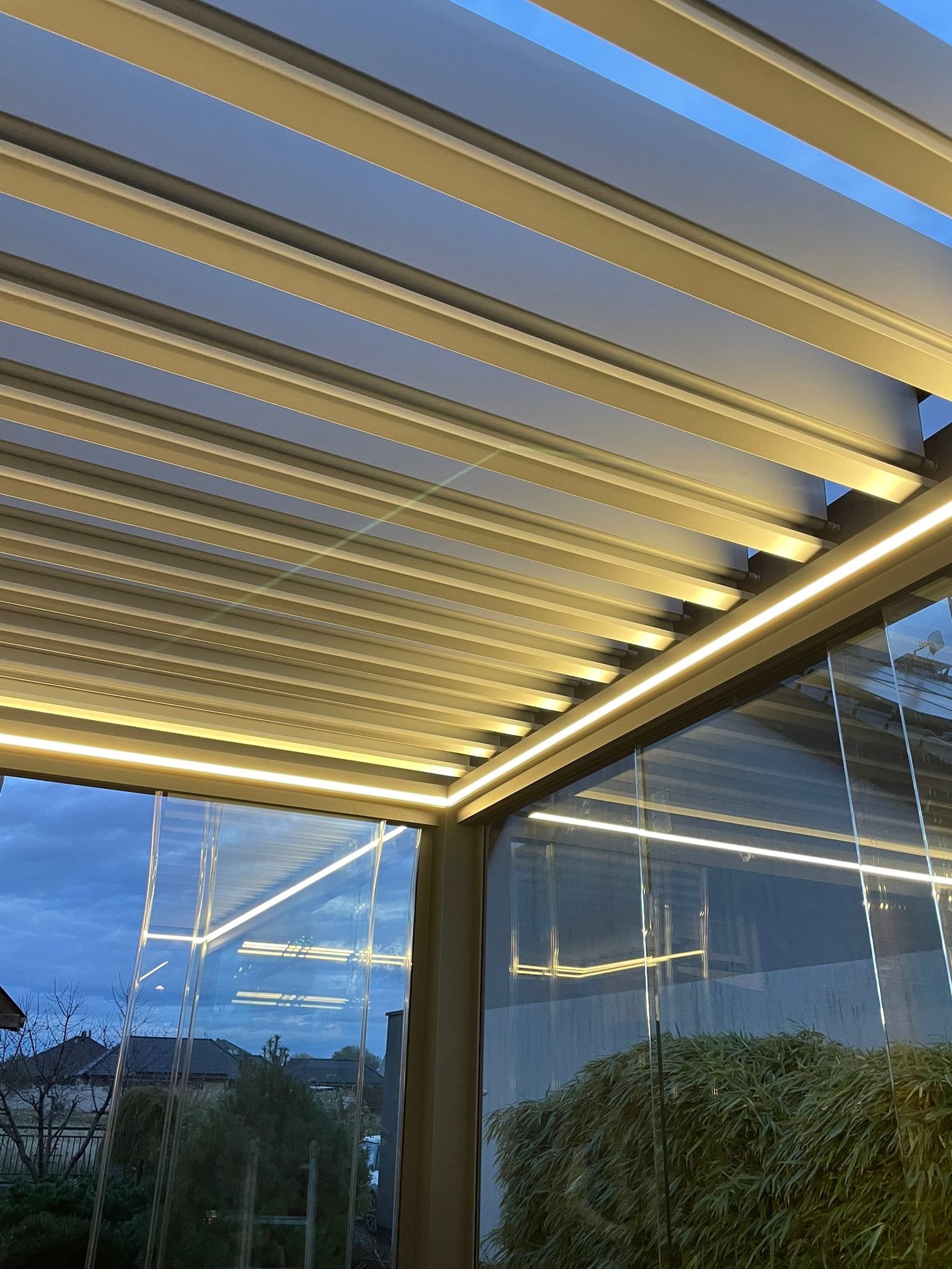 Nowoczesna pergola metalowa z oświetleniem LED, widok z dołu na lamelowy dach i szklane ściany. Wieczorny krajobraz odbija się w szybach.