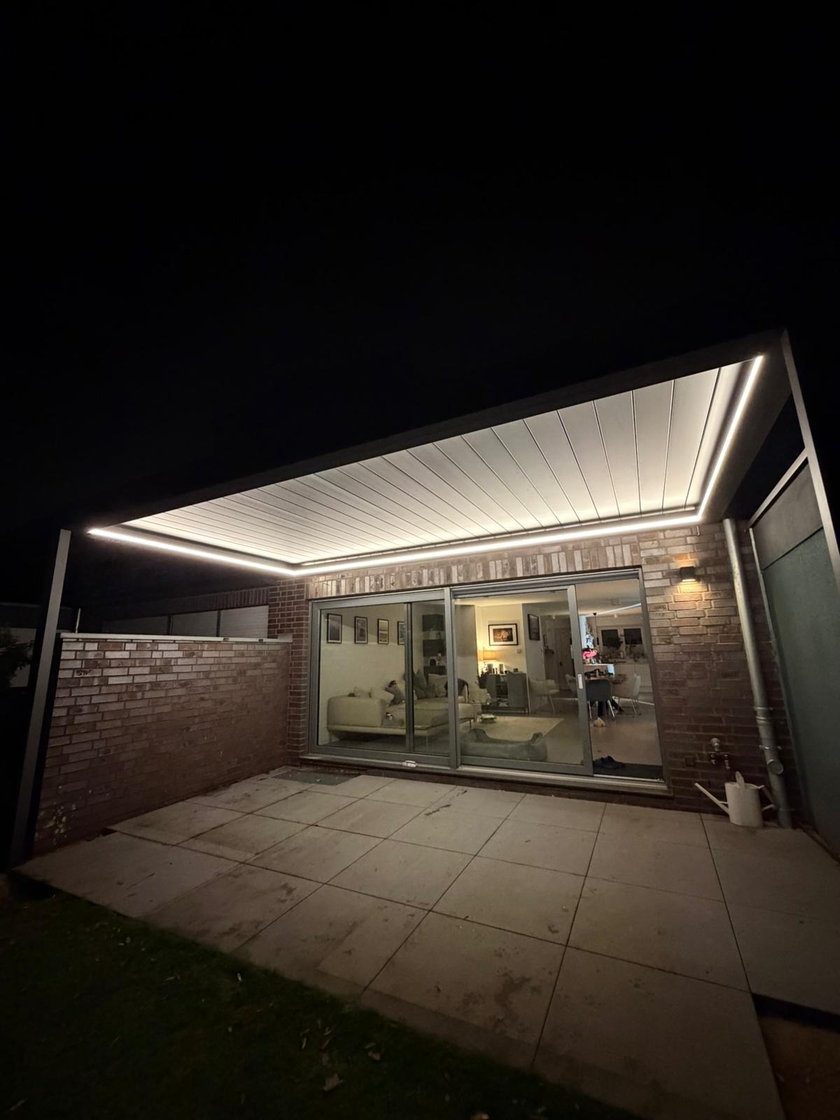 Nowoczesna pergola metalowa z oświetleniem LED, przylegająca do budynku z ceglaną ścianą i przesuwnymi drzwiami tarasowymi, widok nocą.