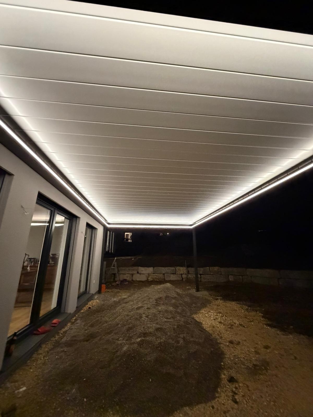 Nowoczesna, podświetlana pergola metalowa z poziomymi panelami, oświetleniem LED i widoczną konstrukcją wsporczą, oświetlona w nocy, z widokiem na teren.