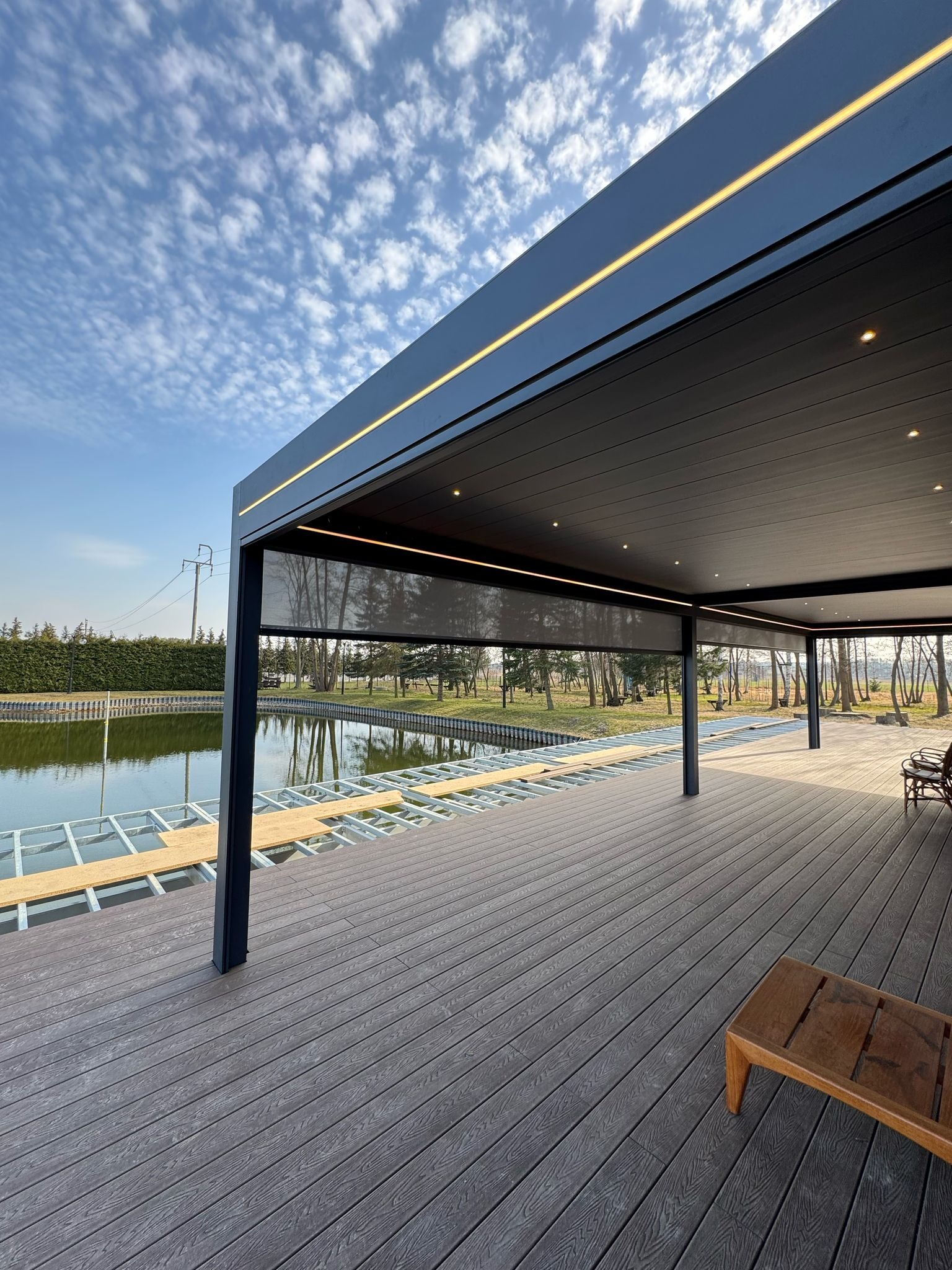 Nowoczesna pergola metalowa z oświetleniem LED i roletami, widok na staw i drewniany taras w trakcie budowy. Błękitne niebo z chmurami w tle.