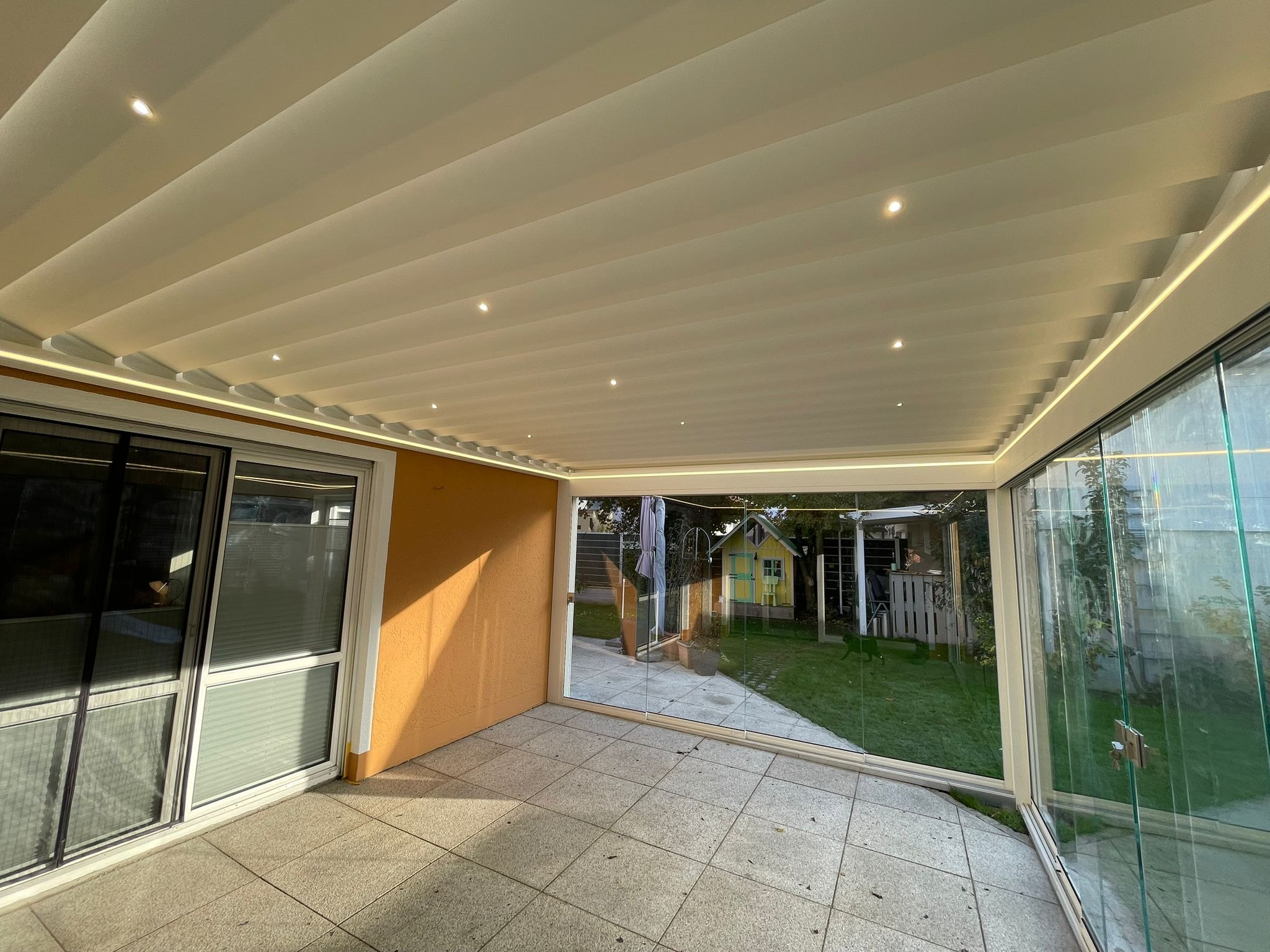 Zadaszony taras z przesuwnymi szklanymi ścianami i oświetleniem LED. Widok na ogród z małym domkiem. Nowoczesna pergola z regulowanymi lamelami.