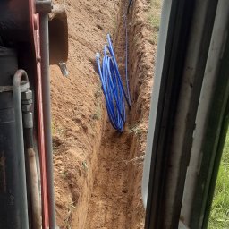 Usługi Koparką Grzegorz Gliwa - Wykopany rów z niebieskimi rurami w ziemi, widok z kabiny koparki. W tle pracownicy i turbiny wiatrowe. Prace ziemne w toku.