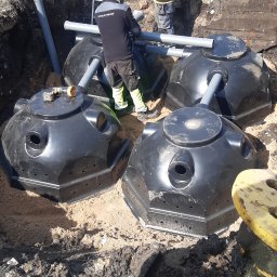 Usługi Koparką Grzegorz Gliwa - Montaż przydomowej oczyszczalni ścieków: cztery czarne zbiorniki w wykopie, pracownicy łączą rury. Widoczny fragment koparki i drabina.