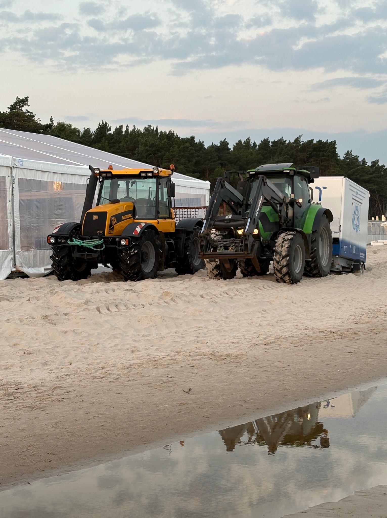 Dwa traktory, żółty JCB Fastrac i zielony z przyczepą toalet, stoją na piaszczystej plaży obok białego namiotu, z lasem w tle i odbiciem w wodzie.